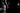 Woman in a white dress standing in a dimly lit room with a man partially visible.