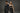 Two people in black outfits standing against a gray background