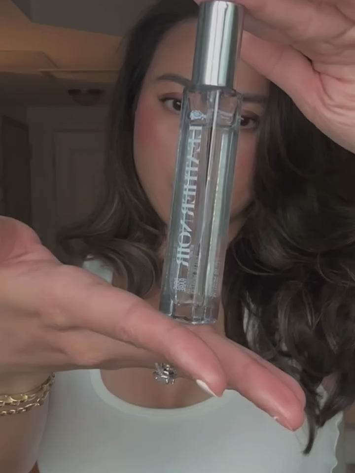 Faith with long, wavy dark brown hair wearing a white tank top, holding a silver cosmetic leather noir travel spray eau de parfum in a home interior setting.