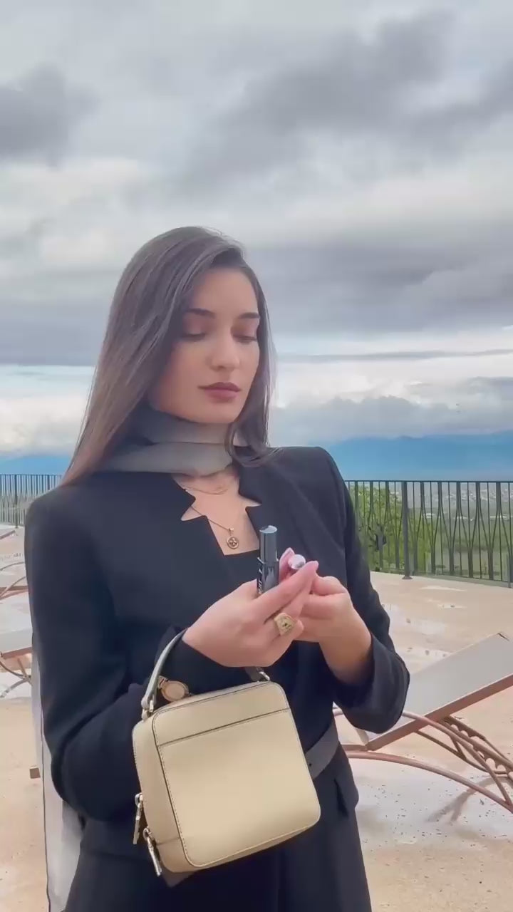Xatia in a black outfit with a beige handbag pointing towards the camera, with a scenic background applying travel spray adore and trackside perfume by petra living