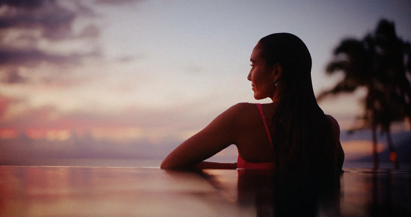 A woman in a pink bikini top sits at the edge of an infinity pool overlooking a sunset over the ocean, with palm trees silhouetted in the background.