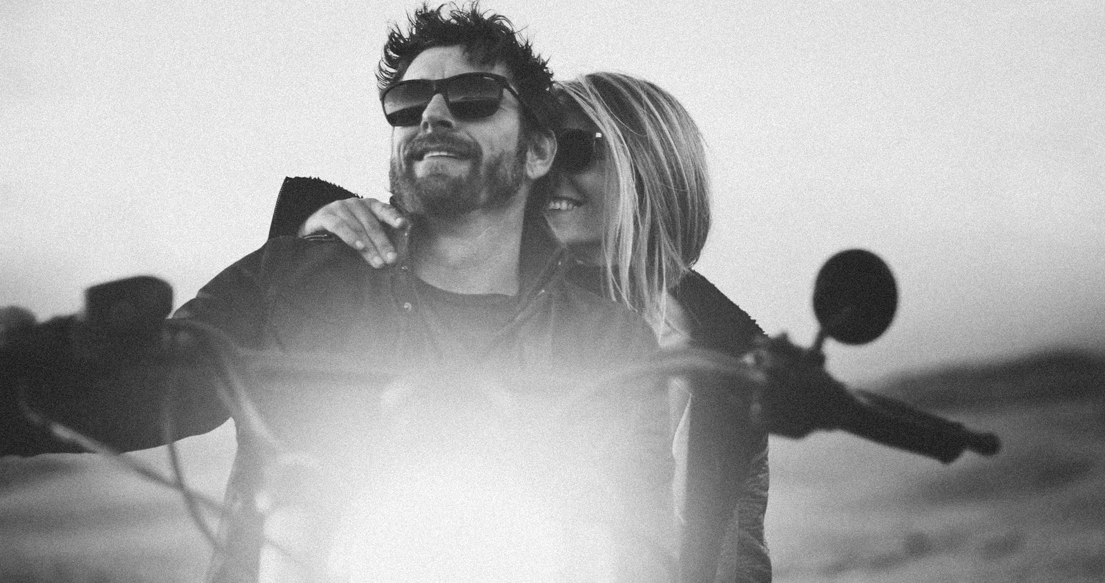 A black and white lifestyle photograph of a couple riding a motorcycle together, with the man in front wearing sunglasses and the woman leaning into his neck.