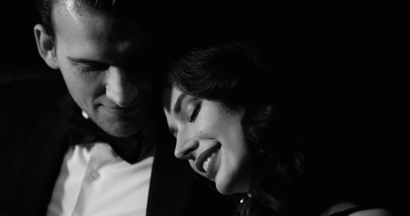 Black and white close-up portrait of a man and woman in formal evening wear, with the man wearing a tuxedo jacket and bow tie and the woman smiling near his face against a dark background.
