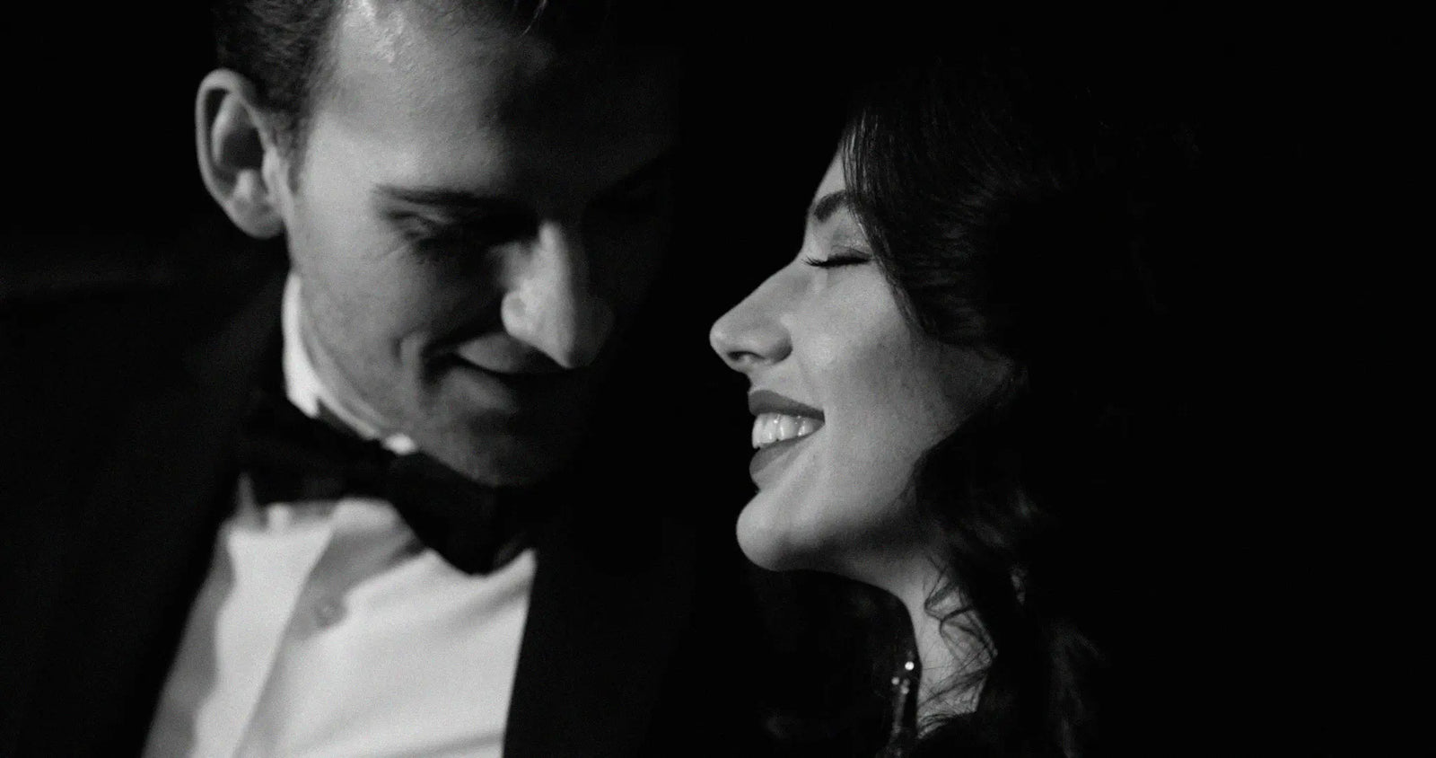 Black and white close-up portrait of a smiling woman looking up at a man in a tuxedo with a bow tie, set against a dark background.